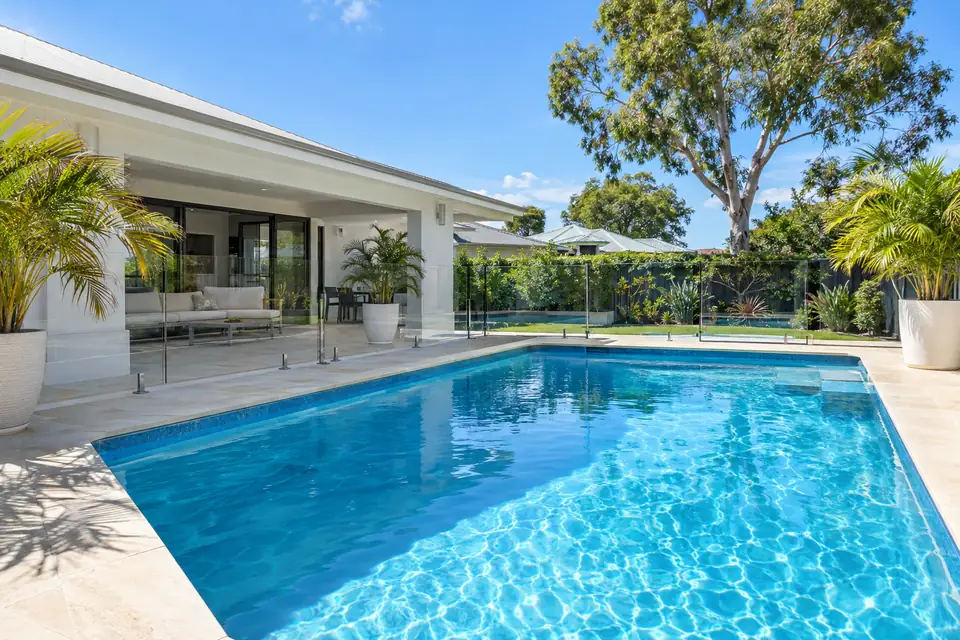 Clear blue pool with limestone paving and a glass fence beside a white rendered home