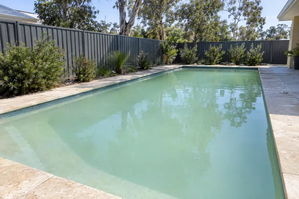 Pool with cloudy, pale green-blue water and limestone paving beside a grey fence
